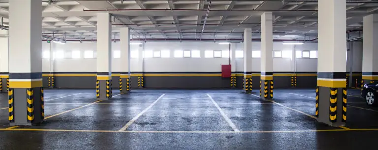 Spacious underground parking garage with painted columns and marked parking bays. Bright natural light filters through windows, highlighting the clean floor
