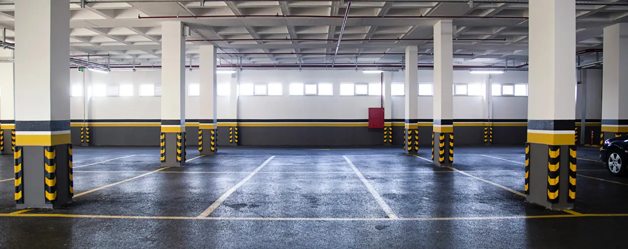 Spacious underground parking garage with painted columns and marked parking bays. Bright natural light filters through windows, highlighting the clean floor
