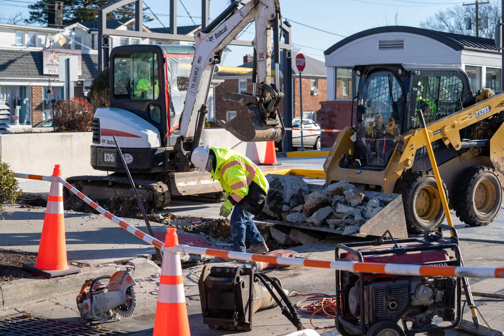 Construction workers using excavators and loaders to remove pavement at a job site, marked with cones and tape for safety during utility repairs