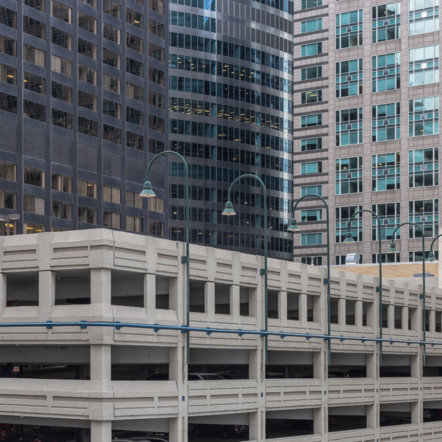 Concrete parking structure surrounded by modern glass and stone high-rise office buildings in a dense urban setting with overhead street lamps