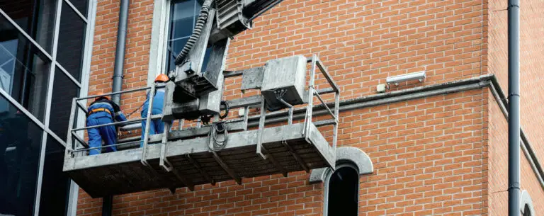 Two construction workers wearing safety gear perform maintenance on a brick building façade while standing on a suspended aerial work platform