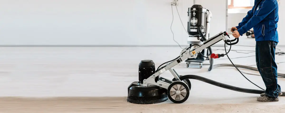 A construction worker uses a concrete grinder indoors to smooth and prepare a floor, with a connected vacuum system for dust control in the background