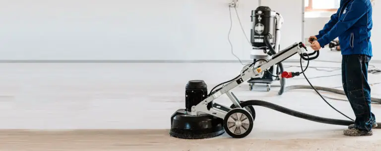 A construction worker uses a concrete grinder indoors to smooth and prepare a floor, with a connected vacuum system for dust control in the background