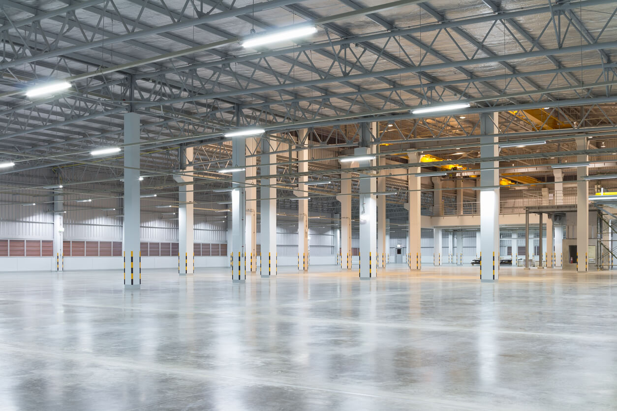 A large, clean warehouse featuring a smooth, polished concrete floor with bright overhead lighting and high ceilings with exposed beams