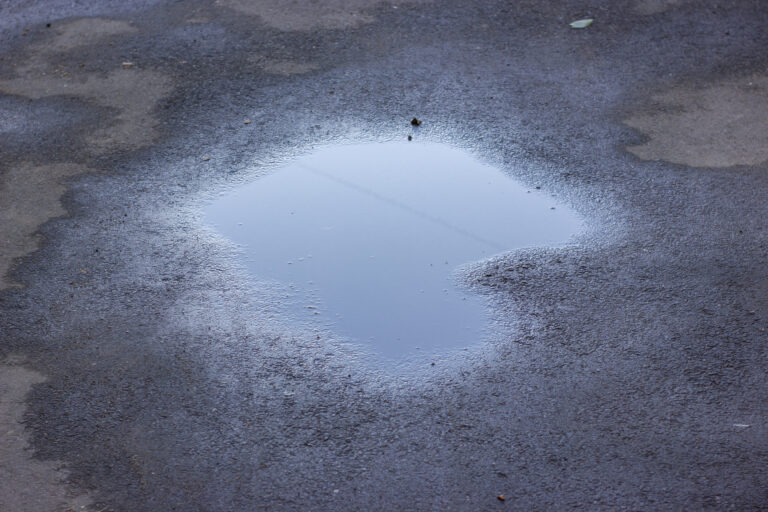 Close-up of a reflective water puddle on dark, fresh asphalt. The pavement is slightly worn with visible patches and a moody, wet texture