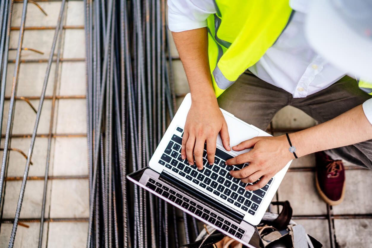 A construction professional in safety gear working on a laptop, reviewing structural plans beside rebar on a job site