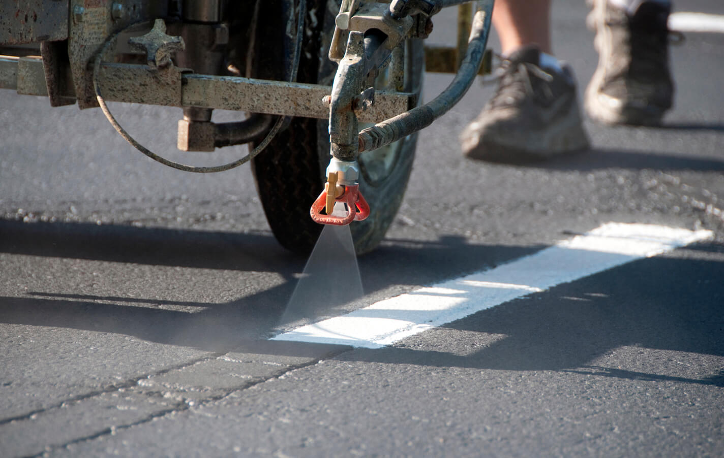 Close-up of a line striping machine applying fresh white paint to asphalt, with a worker nearby during pavement marking work