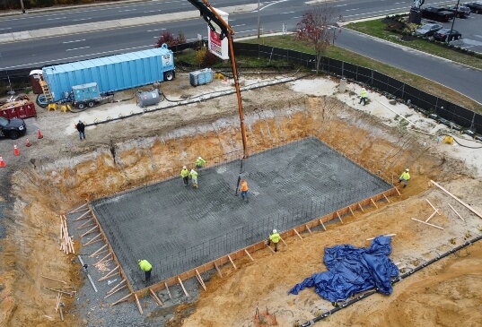 Workers pour and smooth concrete into a rebar-reinforced foundation pit at a commercial site near a roadway, using a concrete pump