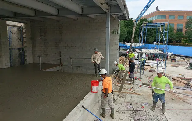 Crew members guide and smooth concrete during slab pour with pump truck support at a partially enclosed commercial construction site
