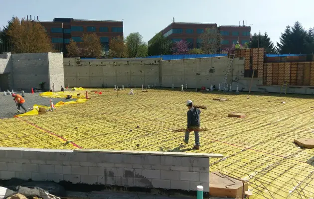 Workers installing rebar grid and vapor barrier across a large foundation slab area in preparation for concrete pour at a commercial site