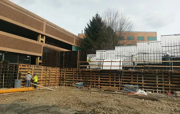 Construction crew assembling wall formwork and vertical rebar adjacent to a parking structure and building during early wall phase