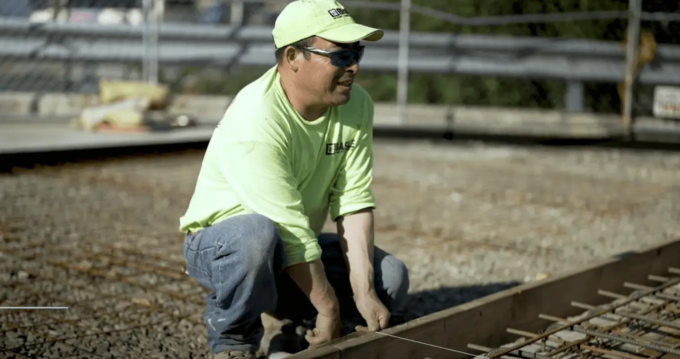 Wearing a neon green shirt and protective glasses, a concrete worker prepares a form with rebar reinforcement on a gravel base