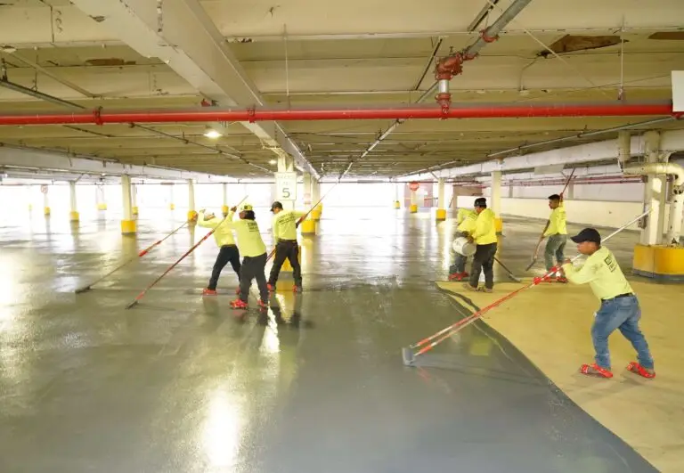 Crew members apply protective coating to a concrete parking garage floor using rollers and squeegees, preparing the surface for long-term durability
