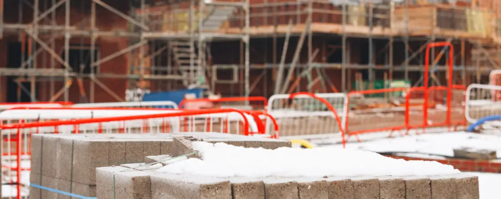 Snow-covered cinder blocks in foreground of job site with safety barriers and structure under renovation